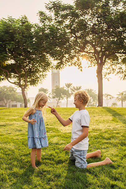 Two children enjoying their time outdoors in a scenic park, surrounded by greenery under the sunlight. The atmosphere is cheerful, with one child playfully engaging with the other in the foreground. Trees and soft light in the background add charm to the moment.