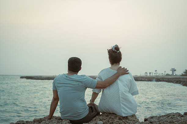 Man and woman sit on seaside rocks, back view, looking at the calm ocean at sunset with copy space—great for travel, romance, mindfulness, lifestyle.