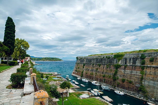 Scenic view of a coastal fortress with boats docked in a calm harbor, surrounded by lush greenery and a cloudy sky.