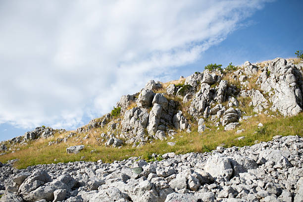 Steep rocky slope with sharp karst formations and scattered grass under a partly cloudy summer sky. Rugged Mediterranean terrain capturing the raw beauty of untouched nature, geology, and remote landscapes.