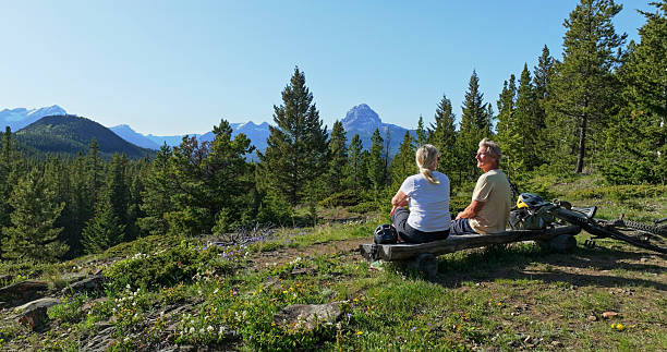Mature couple with e-bicycles relax on bench in mountain meadow, springtime, rear view, Crowsnest Pass, Canadian Rockies