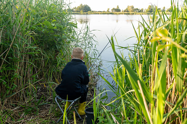 Young boy sitting by riverbank, fishing alone in calm, natural environment on a warm summer day