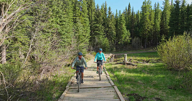 istock Front view of mature couple riding e-bicycles on boardwalk in forest 2225251294