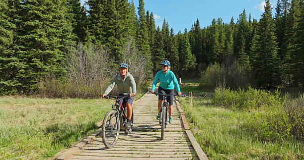 Front view of mature couple riding e-bicycles on boardwalk in forest wilderness area, springtime, Crowsnest Pass, Canadian Rockies