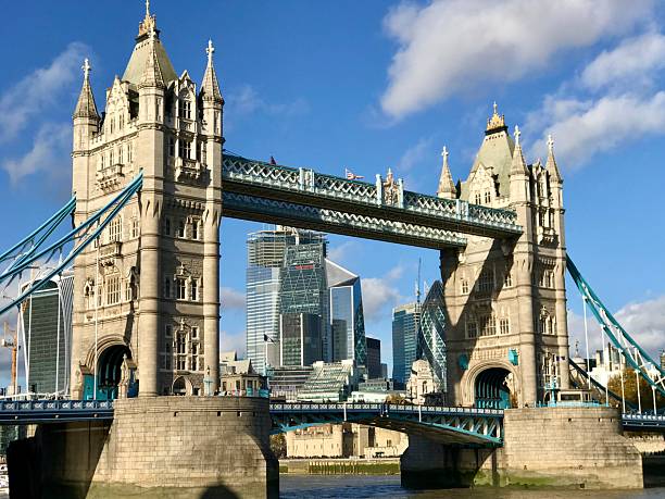 East London, England, The United Kingdom, Great Britain — July 10th 2023. The Cityscape of London. London's Iconic Landmark historic structure the tower bridge which is A Victorian Gothic Revival style structure Captured on a summer day urban view by the River Thames with clear blue sky and white clouds. In the far background, the skyline of Canary Wharf in the London Borough of Tower Hamlets is seen. The east London area.