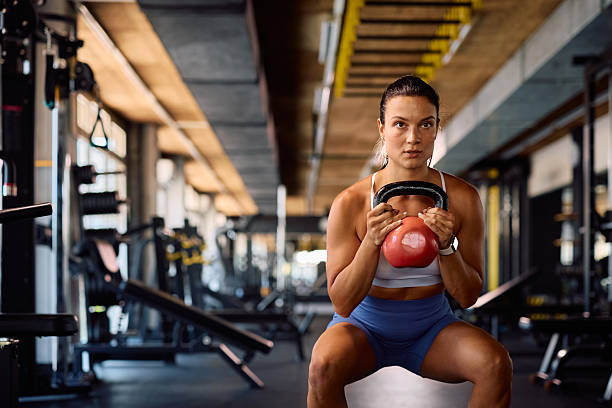 Young sportswoman using kettlebell while doing squats during gym workout. Copy space.