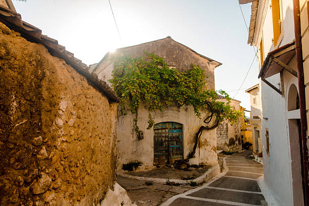 A rustic Mediterranean street with old stone buildings, a vine-covered house, and warm sunlight.