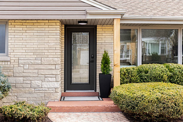 A front door detail with a yellow brick and stone siding, black front door with pattern frosted glass, and a brick sidewalk leading to the red covered concrete porch.