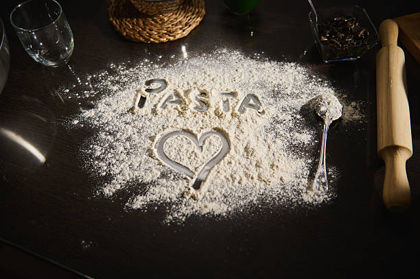 Creative depiction of the word 'PASTA' and a heart symbol drawn in flour on a dark surface, surrounded by baking tools and ingredients, emphasizing culinary art and homemade meal passion.