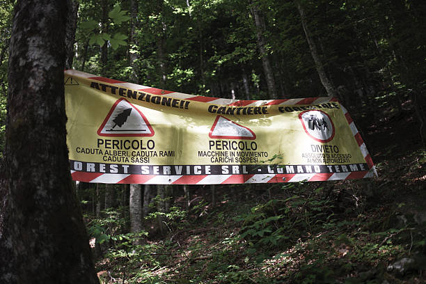 Abetone, Italy - July 10, 2025: A forestry worksite warning banner is displayed between trees in a wooded area. The sign indicates various hazards, including falling trees, moving machinery, and restricted access. The banner is labeled “Cantiere Forestale” and includes safety icons and warnings in Italian, issued by Forest Service SRL - Comano Terme