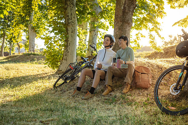 Two friends enjoying a sunny day sitting under trees with bicycles nearby in a peaceful outdoor environment, bonding and relaxing together surrounded by nature, expressing joy, partnership, and tranquility.