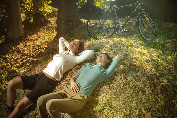 Two individuals enjoy a peaceful rest on the grass with bicycles nearby under the soft sunlight, surrounded by trees and nature, evoking a sense of friendship, tranquility, and connection with the outdoors.