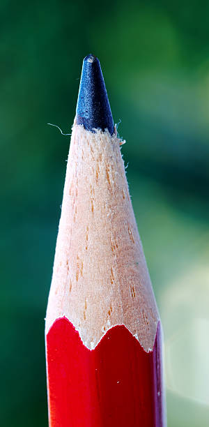 Close-up macro photograph showing the detailed tip of a traditional red pencil, beautifully sharpened, with its wooden grains and black lead visible. The green blurred background creates an artistic, contrasting atmosphere.