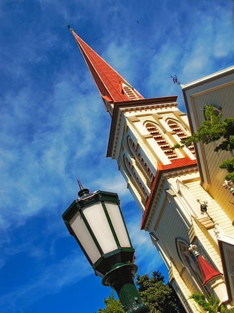 An aesthetic perspective showcasing a tall church spire accompanied by a vintage lantern under a bright blue sky, surrounded by lush foliage. Architectural details are highlighted, emphasizing cultural and historical significance.