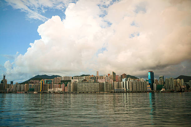 A scenic view of the Hong Kong waterfront with a dense skyline of residential and commercial buildings under dramatic clouds. Taken from across Victoria Harbour, this image captures the unique blend of urban density and natural beauty.