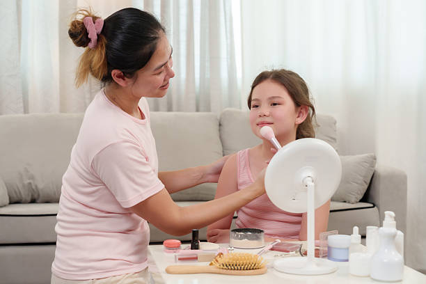 istock Asian single mother applying makeup foundation with brush on young daughter's cheeks, sitting together at cozy home, mother teaching beauty basics, loving connection, emotional parenting 2225297235