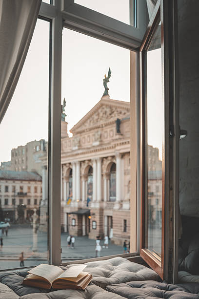 istock Cozy reading nook with an open book on a cushioned window seat overlooking a historic European city square and opera house at sunset through soft curtains 2225314774