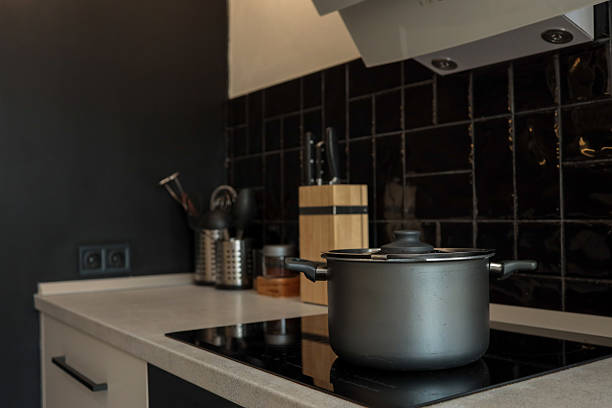 Modern kitchen interior with a matte black cooking pot on a sleek induction cooktop, surrounded by black tiles, a knife block, and metal utensil holders.