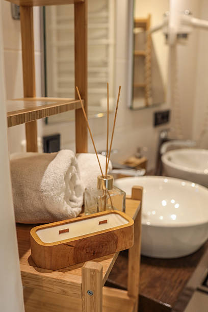 istock Cozy bathroom scene with a rolled white towel, aroma diffuser, and a handcrafted wooden candle on a bamboo shelf near modern ceramic sinks 2225316420