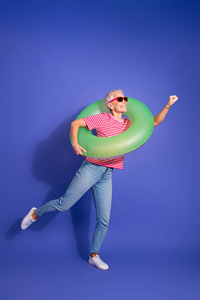 istock Confident elderly woman with green inflatable ring posing against blue vertical background showcasing cheerful summer vibes 2225317488