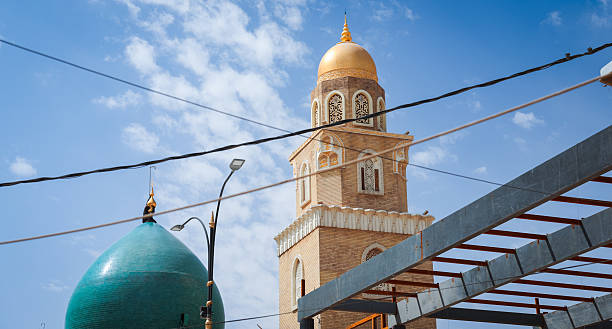 A beautiful mosque tower with a golden dome stands prominently in Kufa, Iraq. The clear blue sky provides a vivid backdrop.