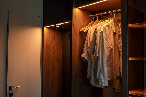 istock Modern wooden wardrobe with LED lighting and several light shirts on hangers. Minimalist interior design with open closet and contemporary door 2225319306