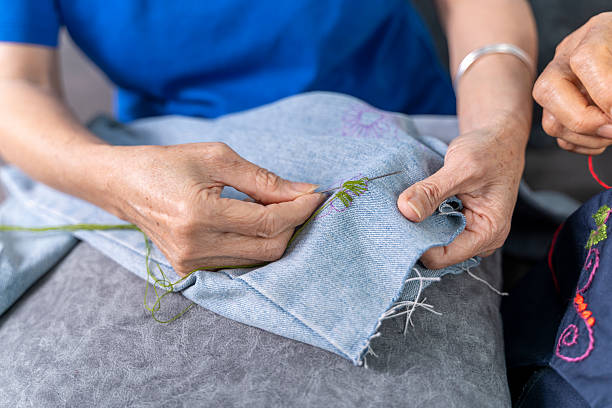 Close-up of elderly women's hands hand-stitching green thread embroidery on denim, showcasing traditional textile craftsmanship and creativity.