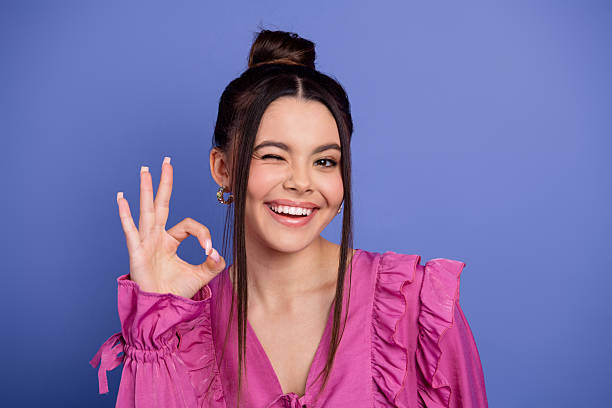 istock Charming young woman winking while showing an OK gesture against a purple background, featuring trendy fashion and style 2225320643