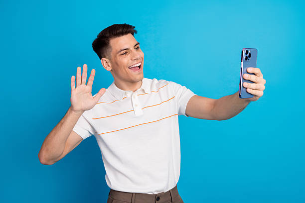 istock Joyful young man waving at smartphone camera on blue backdrop, showcasing casual attire for creative projects 2225320979