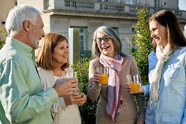 Waist up view of man and three women, all in their 60s, casually dressed, standing in sunshine of rooftop garden, talking and laughing.