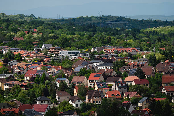 A vibrant suburban neighborhood with numerous detached houses, many equipped with solar panels on their red or brown tile roofs. Surrounded by dense trees and rolling hills in the background, the area presents a harmonious blend of sustainability, modern living, and traditional European architecture.