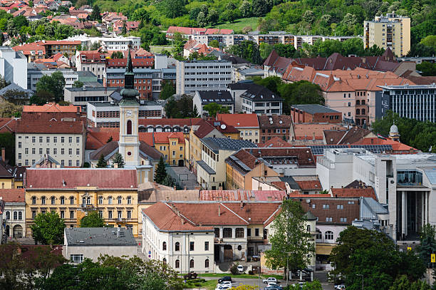 A detailed aerial view of a European town center showcasing a mix of architectural styles, from ornate historic buildings with red-tiled roofs to modern office structures. The clock tower stands as a focal point among the colorful facades, all nestled in a green urban landscape.