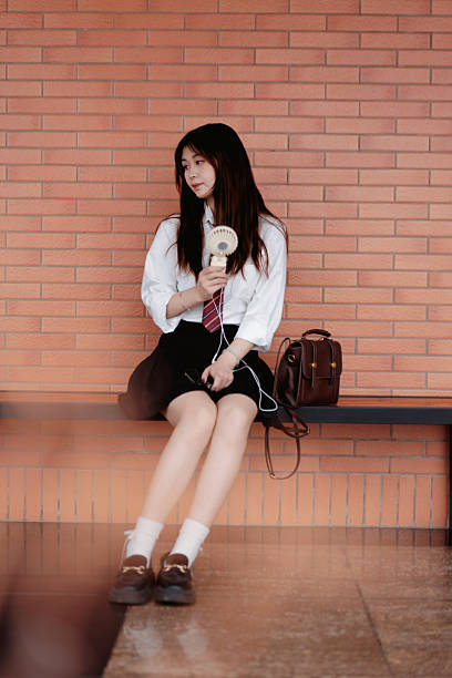 Young Asian female student in school uniform sitting on a bench against a red brick wall, listening music through earphone and holding an electric mini fan.