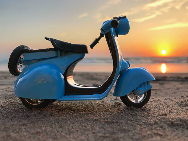 A blue scooter is sitting on the sand. The sun is setting in the background. The scooter is on the beach
