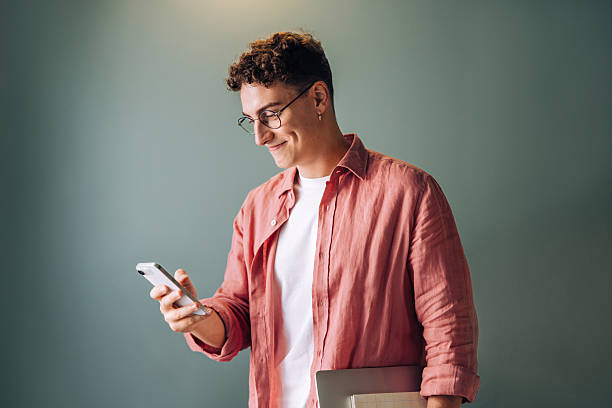 young man using smartphone while holding a laptop and smiling - endast en ung man bildbanksfoton och bilder