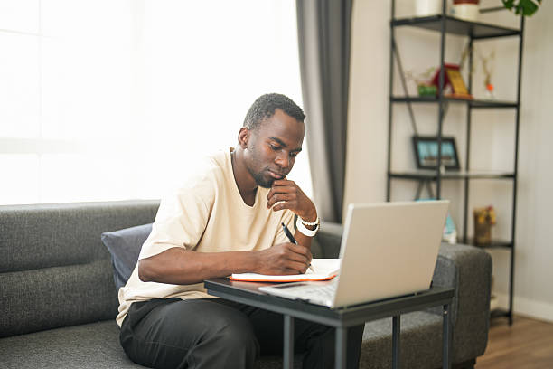 istock Young African man working remotely from his living room, writing in a notebook 2225353574