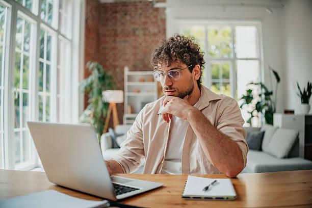 istock Young man focused on his laptop while working from home in a modern living room filled with natural light and greenery 2225356112