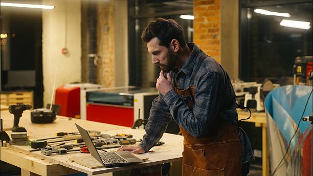 istock Cheerful Caucasian man wood master carpenter using laptop in workshop builder searching information in Internet guy male chatting social media network pc repairman smile workman order materials online 2225363468