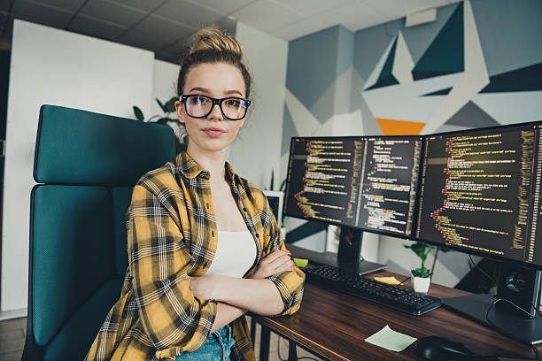 Young female programmer working in a casual environment with a dual-monitor desktop setup, featuring coding screens