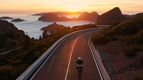 Epic Sunset View in High Mountain Landscape with Motorcycle Driver Riding on Asphalt Road . Pico do Arieiro , Madeira Island . Clouds in the Valley .