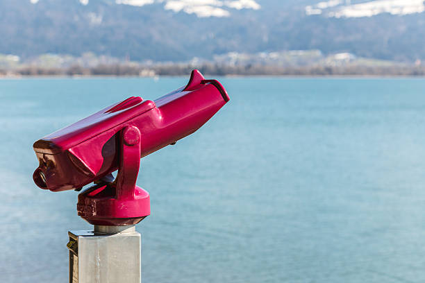 A red telescope is pointed at the water. The water is blue and calm. The scene is peaceful and serene
