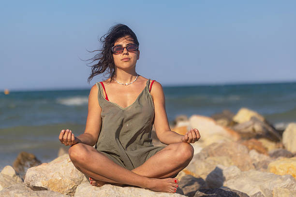 A dark-haired woman in a relaxed green beach cover-up sits peacefully meditating on the rocks by the sea. With her eyes closed and hands in a meditative pose, she appears deeply immersed, finding serenity in the rhythmic sound of the waves.
