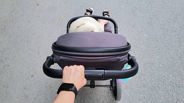 Top-down view of a parent pushing a modern baby stroller on an asphalt path. A child in a straw hat is sitting in the stroller, with their legs visible. The parent's hand, wearing a smartwatch, holds the stroller handle.