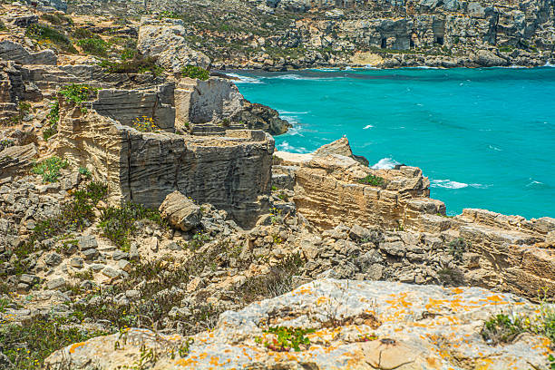 Ancient limestone area on Favignana Island, Sicily, with rugged rock formations, sparse vegetation, and a deep blue sea under a bright sky in the background