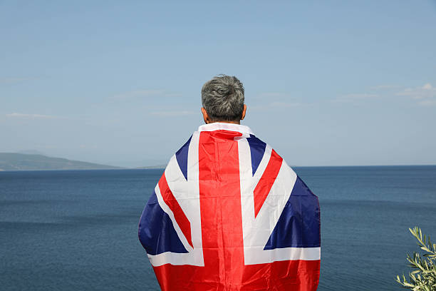 Proud senior man wrapped in the Union Jack flag, raising his arms in triumph while standing by the calm sea. With gray hair and a strong stance, he radiates patriotism, strength, and connection to national identity. The backdrop of open water symbolizes freedom, pride, and belonging.