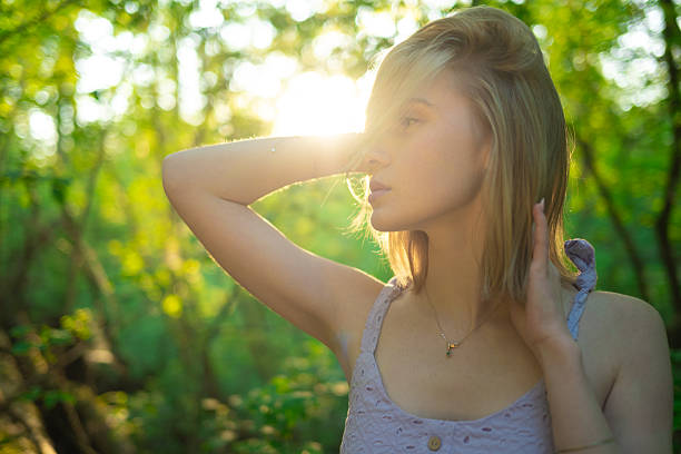 Portrait young blonde woman in a lavender dress posing outdoors in soft sunlight, surrounded by green foliage.