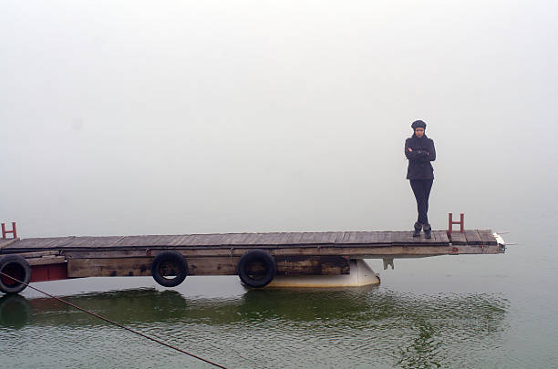 Young sad lonely woman standing on the river dock on a misty autumn day.