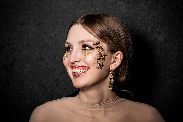 A woman with brown hair and gold pieces makeup wears a sheer beige top and gold earrings. She stands against a textured black background, showing her neck and collarbone.