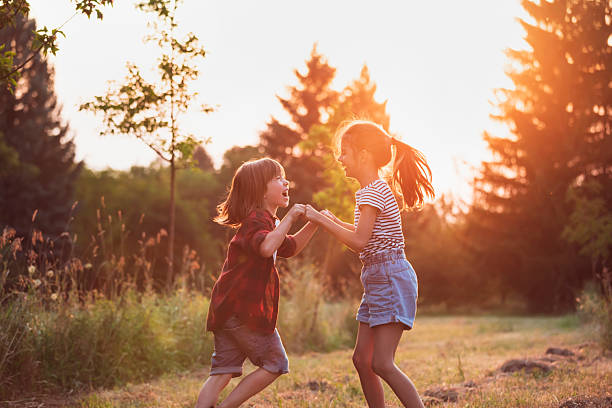 Cheerful boy and girl spinning around while holding hands outdoors in sunset light.