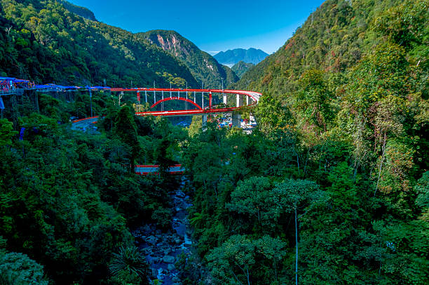 Beautiful mountain scenery with a striking red bridge known as Kelok 9 (Kelok Sembilan) in West Sumatra, Indonesia. The bridge winds gracefully through lush green hills and stretches over a deep valley with a river flowing below.